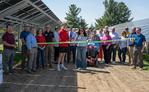Group of people at a ribbon-cutting ceremony in front of solar panels outdoors, including someone using a mobility device.